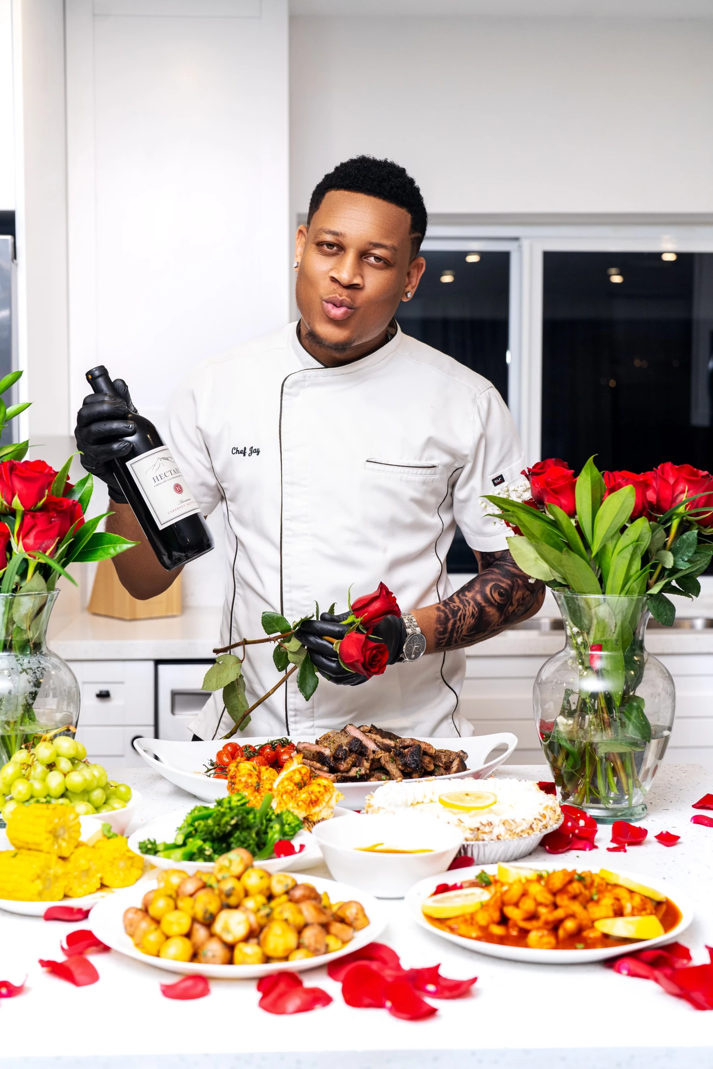 Chef Jay holding a wine bottle and a rose behind a dining spread in his kitchen