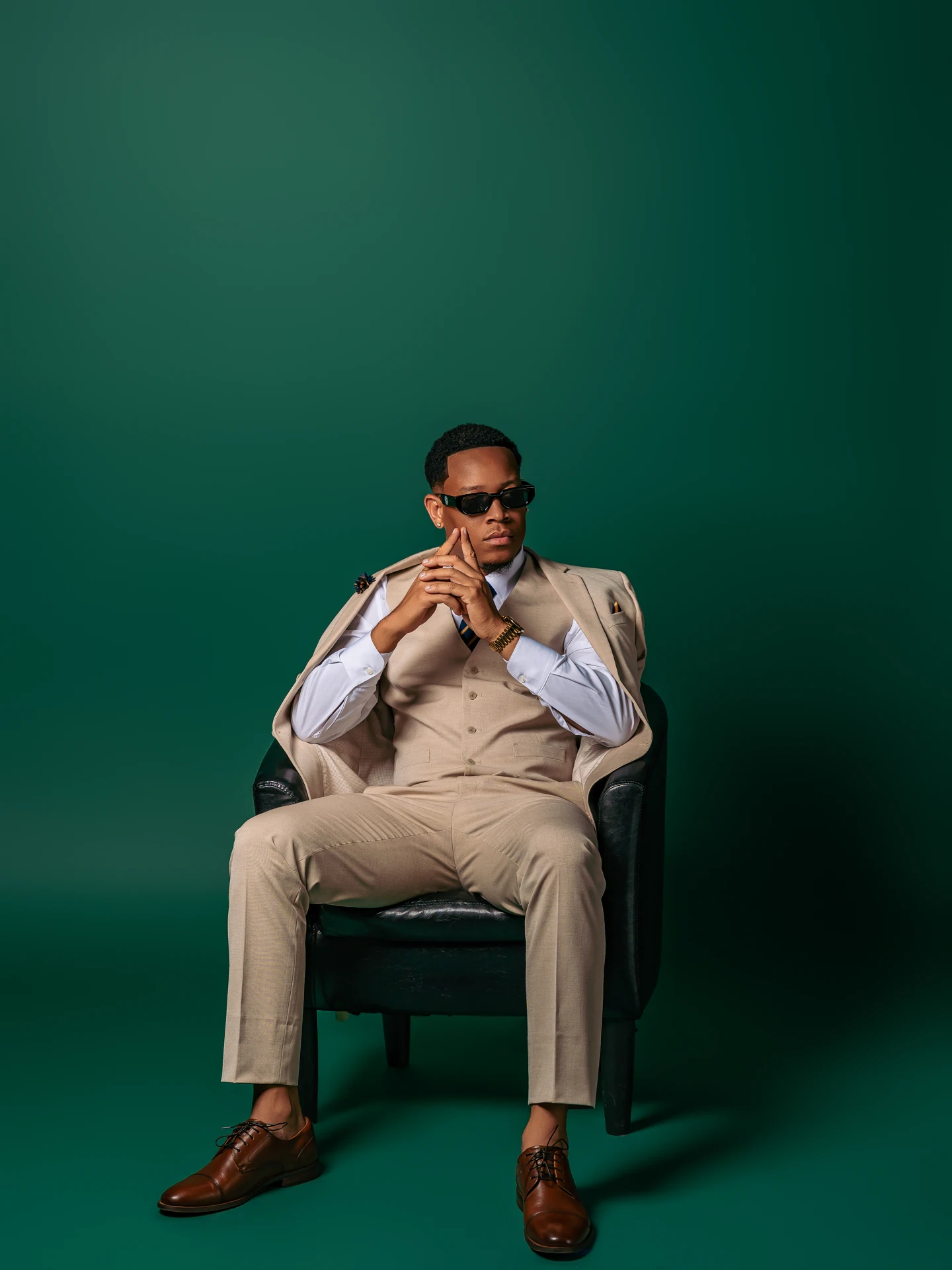 Jemar Lyon seated in a beige waistcoat and trousers with his hands clasped against a deep green studio backdrop