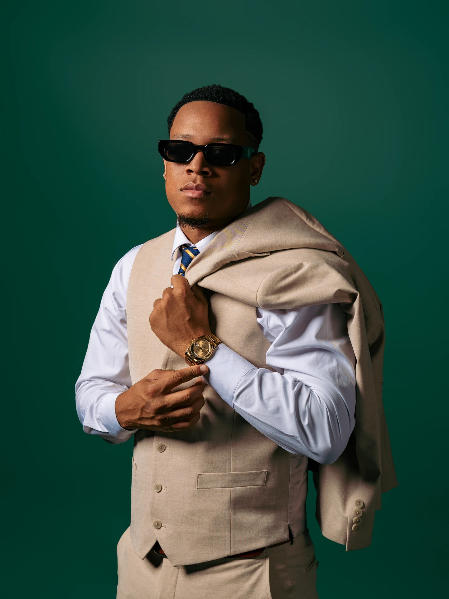 Jemar Lyon in sunglasses and a tailored beige waistcoat, with his jacket draped over his shoulder against a deep green backdrop