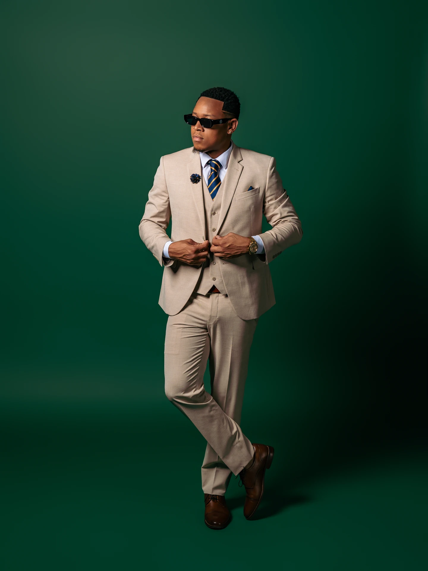 Jemar Lyon standing in a tailored beige three-piece suit and sunglasses against a green studio backdrop